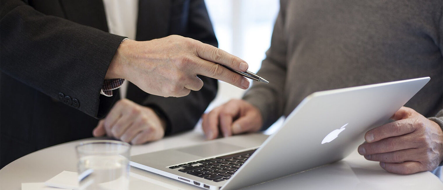 ausgestreckte Hand zeigt mit einem Stift auf den Bildschirm eines aufgeklappten Laptops, zweite Person steht daneben ausgestreckte Hand zeigt mit einem Stift auf den Bildschirm eines aufgeklappten Laptops, zweite Person steht daneben
