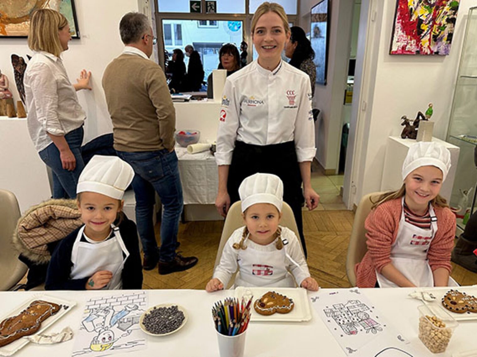 Drei Kinder sitzen am Tisch, im Hintergrund Rowena Redwanz Drei Kinder sitzen am Tisch, im Hintergrund Rowena Redwanz