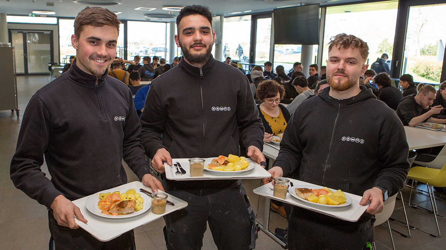 Drei Auszubildende präsentieren in der Mensa ihre ausgewählten, gesunden Mittagessen.